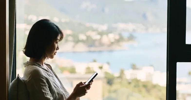 Traveler planning a trip on their phone by a window overlooking a coastal bay